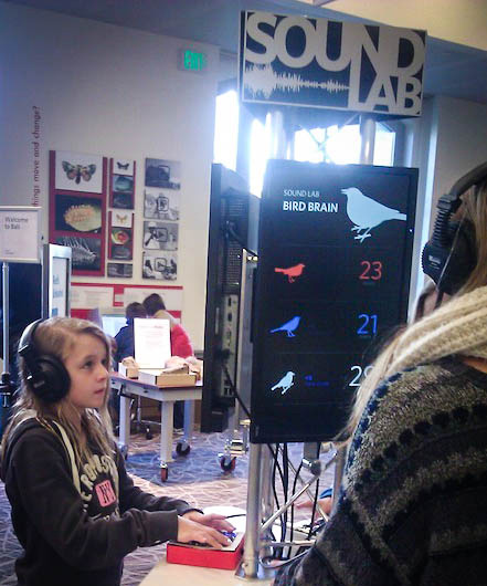 A child in headphones at the Sound Lab cart, hand on the arcade buttons, scoreboard on the screen above